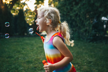 Kid child  trying to catch eat soap bubbles with tongue. Candid portrait of cute funny little blond Caucasian girl playing on home backyard at summer sunset. Real authentic happy childhood moment.