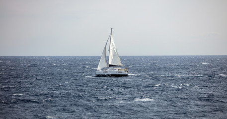 Sailboat travels with wind help, against the waves. Wavy sea, blue sky background.