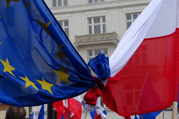 Flaga Unii Europejskiej i Polski splecione, związane razem, podczas demonstracji poparcia dla członkostwa Polski we Wspólnocie, w tle, rozmyte, budynki, inne flagi, głowa mężczyzny, z tyłu - obrazy, fototapety, plakaty