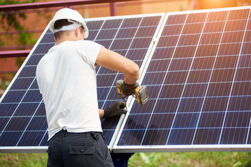 Back view of young technician connecting solar photo voltaic panel to metal platform using...