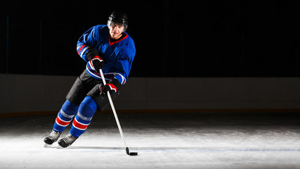 Naklejka premium Young hockey player with stick and puck skating on rink in attack against dark background