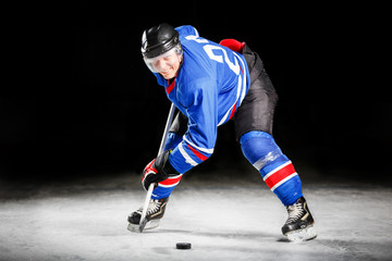 Young hockey player with stick and puck skating on rink in attack against dark background