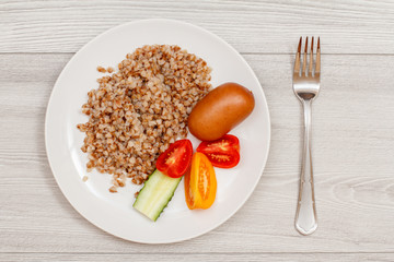 Buckwheat porridge with sausage and slices of tomato and cucumber on white plate