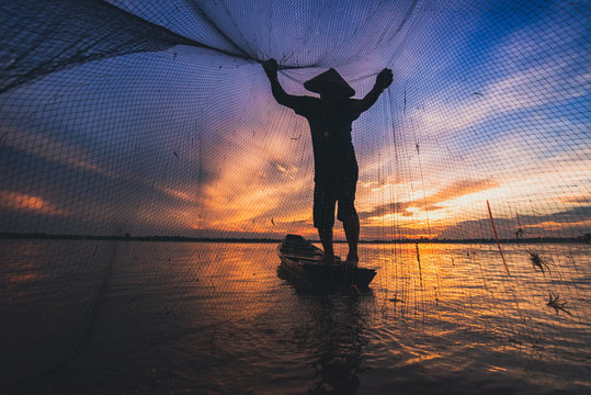 Asian fisherman on wooden boat casting a net for catching freshwater fish in nature