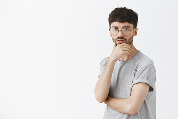 Creative and artistic male designer in round glasses and beard touching chin and gazing thoughtfully at camera, thinking or making up ideas for new fashion week clothing design over gray wall
