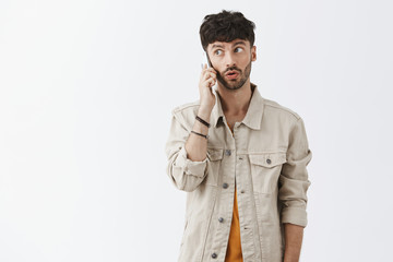 Man feeling intrigued hearing interesting offer via smartphone listening carefully while talking on cellphone gazing left posing with phone near ear and folded lips over gray background