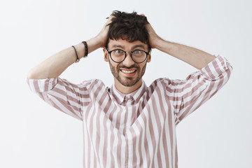 Waist-up shot of creative confused and worried good-looking male artist in glasses and stylish striped shirt holding hair with hands and smiling intense feeling worried with lack of time