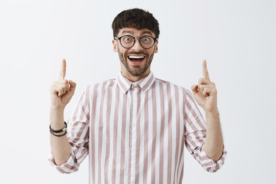 Excited good-looking happy man with beard and dark cool hairstyle in black glasses and striped shirt raising index fingers pointing up with amazed broad smile, feeling impressed and astonished