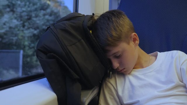 The boy is traveling by train sleeping near the window on his backpack. Sleep after a hard day traveler