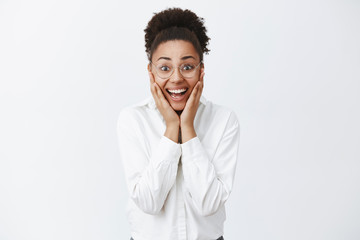 Fototapeta na wymiar Dream come true. Surprised happy and joyful attractive female entrepreneur in glasses and white shirt with dark skin, holding hands on cheeks and smiling broadly from amazement and happiness