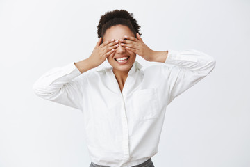 Indoor shot of charming carefree african-american female employer with white shirt, covering eyes with palms, being blinded while playing peek-a-boo and smiling broadly at camera over grey wall