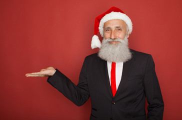 Fototapeta na wymiar Senior man in santa hat an suit presenting something on a red wall, smiling for the camera. Xmas concept.