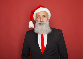 Senior man in santa hat an suit presenting something on a red wall, smiling for the camera. Xmas concept.