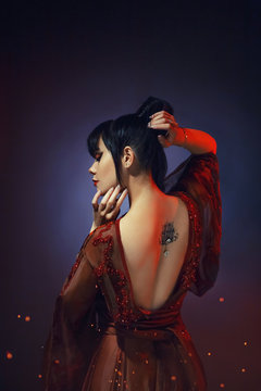 young girl with dark blue hair and a bang in a long red dress with open naked bare back. a tatoo picture lotus with a shiny piercing. hands near the face and hairdo. gloomy background with highlights.