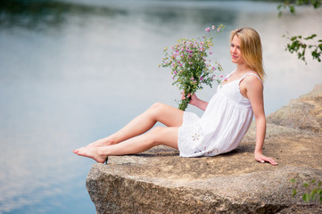 Young woman in white lace dress on the beach. She sits with bouquet of wild flowers in her hands.