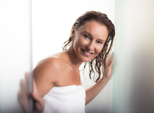 Relaxed and happy. Portrait of beautiful lady with wet hair walking out from bathroom. She is wearing soft towel and looking at camera with smile