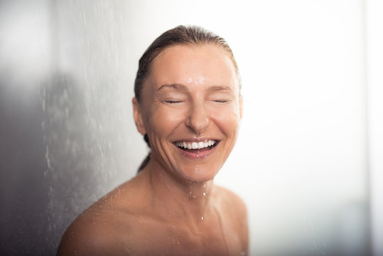Having fun at bathroom. Portrait of attractive lady with closed eyes and wide smile standing under water drops