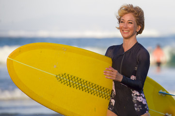 Obraz premium young attractive and happy blonde surfer girl in beautiful beach carrying yellow surf board walking out of the water enjoying summer holidays at tropical island