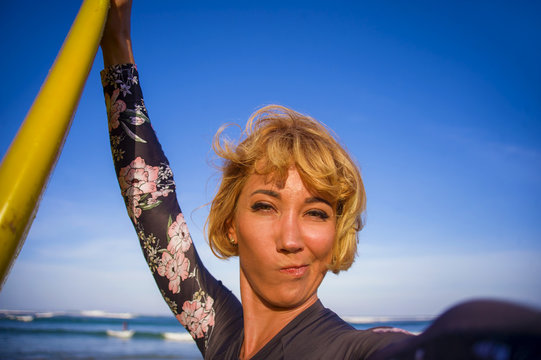 young attractive and happy blonde surfer woman in swimsuit holding surf board in the beach taking self portrait selfie picture smiling cheerful enjoying holidays