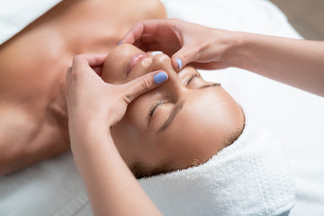 Fototapeta na wymiar Feeling sleepy. Close up portrait of charming woman with closed eyes enjoying skincare procedure at spa salon. Masseuse hands with blue manicure touching client chin and nose