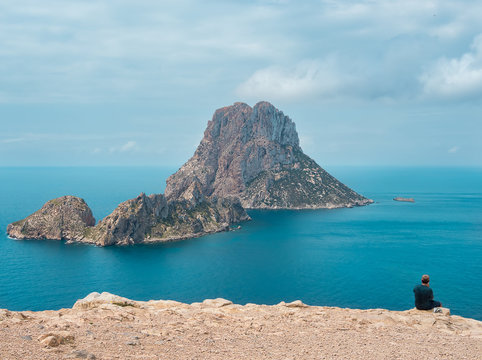 Hombre sentado en acantilado del parque natural de Cala D&acute;hort, observando la belleza de los islotes de Es Vedr&aacute; y Es Vedranell en Ibiza, Espa&ntilde;a.