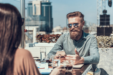 Smiling bearded man telling with woman, while keeping phone in hands. She tasting glass of water while sitting opposite him at table in cafe outside. Glad colleagues communicating during break concept