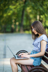 Young woman in eyeglasses sitting on bench in park and using laptop computer