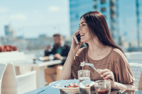 Side view happy girl talking on mobile while tasting appetizing meal with fork outdoor. Man situating at desk opposite her. Glad client using gadget during lunch concept
