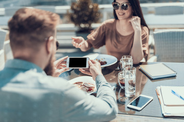 Male talking photo of dish while looking at screen of phone. Positive female talking with him while gesticulating hand. They sitting at desk in cafe during conversation