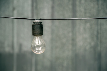 A light bulb on the horizontal wire after the autumn rain. Element of a street garland. In the background a gray iron wall.