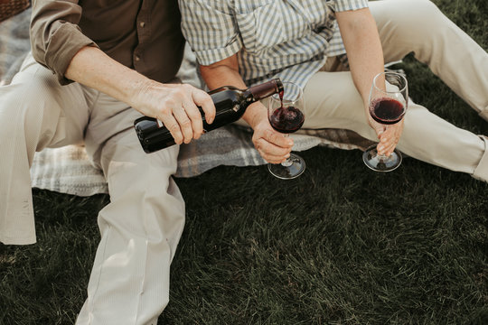 Close up of aged male person pouring wine into glasses his wife hold. They sitting on plaid at green grass. Top view - Powered by Adobe