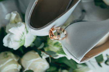 Wedding white bride shoes with a bouquet of white roses and other flowers, wedding rings on a stool.