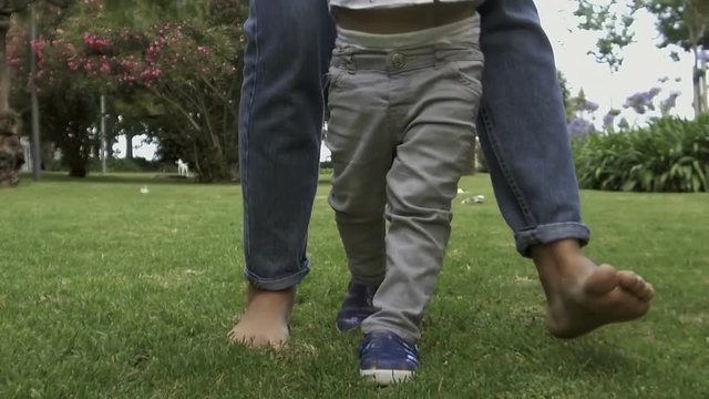 Mother with little son walking on lawn. Barefoot mom teaching cute boy to walk in in slow motion. Low section. Baby boy learning to walk with mother help. Childhood concept
