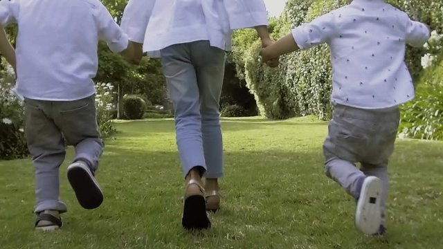 Three kids running to mother together, back view, slow motion. Rear view of children holding hands and running on green lawn in summer. Happy family concept