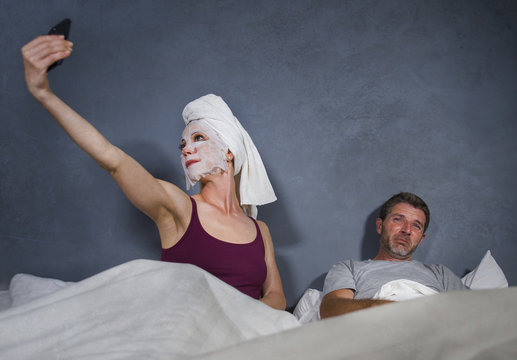 eccentric housewife with makeup facial mask and towel taking selfie in bed and husband with desperate face expression in weird man woman relationship concept