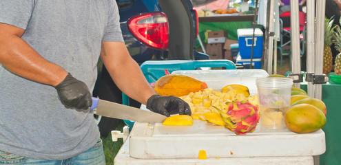 Front view, medium distance of a male, vendor, slicing freshly picked tropical fruit, on display...