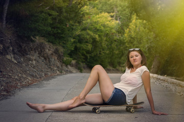 Young woman sits on a skateboard on concrete road. Barefoot relaxed female in jeans shorts and white t-shirt sits with legs stretched out. Small bruise is visible on left leg. Active lifestyle concept