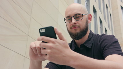 A man with a beard wearing black glasses uses a phone sitting in front of a large building. Close-up shot. Soft focus