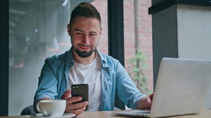 A young man sitting and using a computer and a phone indoors. Medium shot.