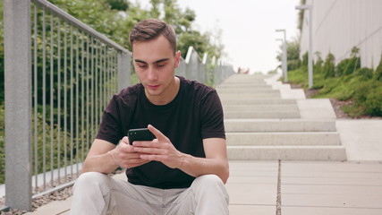 A young man using a phone sitting on the stairs in town. Medium shot. Soft focus
