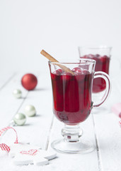 Hot red berry winter drink in two glasses on white wooden table with Christmas tree toys on background.
