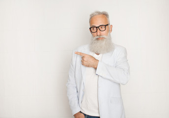 Portrait of old cheerful man in glasses pointing away with finger and smiling over white wall