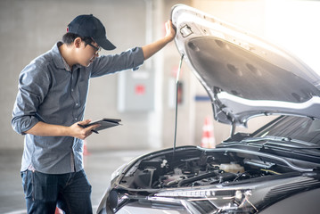 Asian auto mechanic holding digital tablet checking car engine under the hood in auto service...