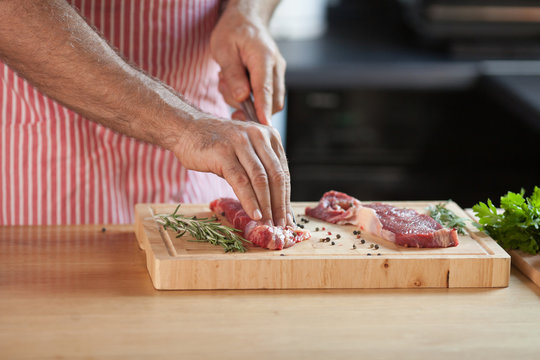 Hands of a man preparing meat and vegetables in a kitchen slicing lean meat on a wooden board.