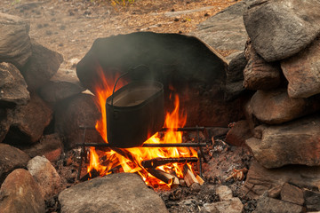 A bonfire in a stone hearth and a tourist cauldron with water. It's about cooking meal at hike.