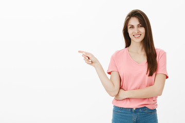 Portrait of beautiful brunette in pink t-shirt smiling joyfully while pointing left with index finger as if inviting to get inside or suggesting visit new store, posing over gray background