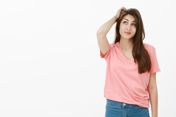 Fototapeta premium Portrait of unsure good-looking carefree female with dreamy expression, smiling and gazing up while scratching head, being clueless, thinking what to answer, standing over grey wall
