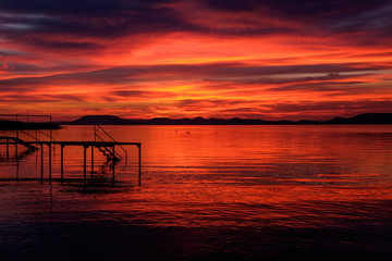silhouette of pier at red and hot sunset at Balaton lake - clouds, thin waves on water and hills in...