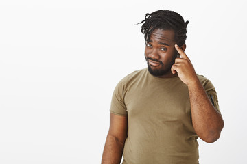 Fototapeta premium Think with head before actions. Portrait of joyful confident african-american with stylish haircut, tilting head and holding index finger on temple while smiling and asking to remember something