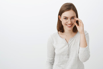 Waist-up shot of joyful creative and smart girlfriend with short brown hair looking from under forehead mysterious and intrigued taking off glasses and smiling broadly having fun over gray wall